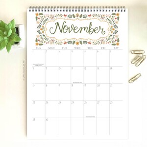 Pu&ograve; includere: Un calendario di novembre a spirale, con un motivo floreale attorno alla parola "Novembre" in corsivo verde. La griglia del calendario bianca ha linee grigie, date, giorni e indicazioni di festivit&agrave;. Include graffette e una piccola pianta.