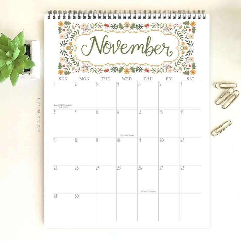 Pu&ograve; includere: Un calendario di novembre a spirale, con un motivo floreale attorno alla parola "Novembre" in corsivo verde. La griglia del calendario bianca ha linee grigie, date, giorni e indicazioni di festivit&agrave;. Include graffette e una piccola pianta.