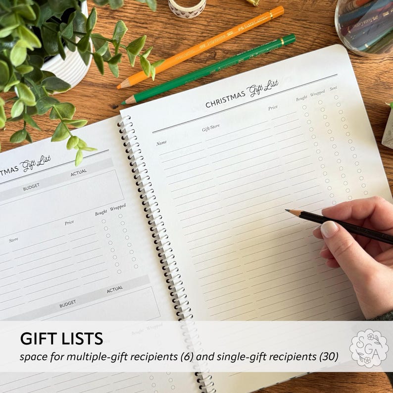 Open Holiday Planner Notebook flat lay showing Christmas Gift List pages for multiple and single recipients. Hand holding pencil ready to write gifts with color pencils beside greenery plants. Holiday gift tracker and Christmas organizer notebook for Christmas shopping and planning.