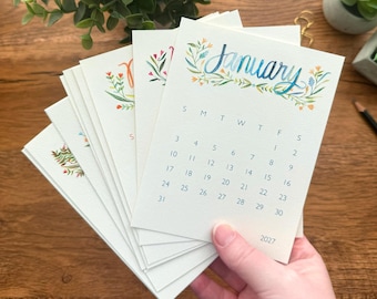 May include: A set of off-white paper calendar cards for the year 2027. The top card displays the month of January in blue cursive script, framed by watercolor floral illustrations. Below the text is a grid of dates. Each card features unique floral art.