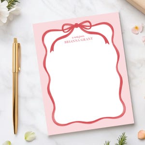 May include: Pink notepad with a red ribbon border and a bow at the top. The text "a note from BRIANNA GRANT" is printed on the notepad.