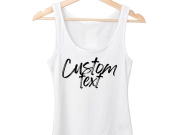 May include: A white tank top with the words "Custom text" in black script. The top is hanging on a wooden hanger against a white background. The tank top has a scoop neck and is sleeveless.