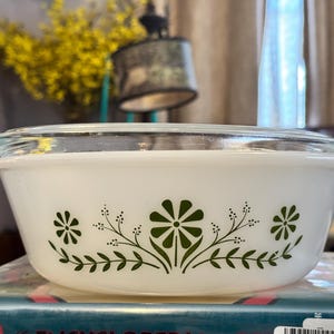 May include: White Pyrex bowl with a clear glass lid. The bowl features a green floral design around the exterior. The bowl is sitting on top of a book.