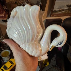 May include: A ceramic swan-shaped planter or decorative vessel featuring an iridescent, pearlescent white glaze. The swan has a curved neck, a detailed wing design with raised feathers, and a gold-painted beak. The surface reflects light.