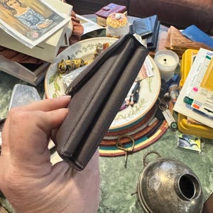 May include: A dark brown leather wallet with a rectangular shape and a clasp closure. The wallet is held in a hand, with the details of the stitching visible along the edges. The background includes a plate, a candle, and other items.