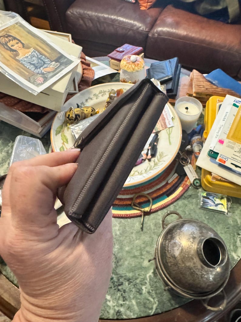 May include: A dark brown leather wallet with a rectangular shape and a clasp closure. The wallet is held in a hand, with the details of the stitching visible along the edges. The background includes a plate, a candle, and other items.