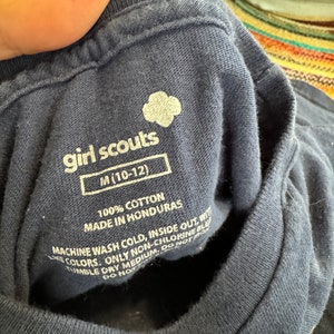 May include: A navy blue Girl Scouts t-shirt with the Girl Scouts logo and the text "girl scouts" and "M (10-12)". The tag also includes care instructions: "100% Cotton, Made in Honduras, Machine Wash Cold, Inside Out, With Like Colors, Only Non-Chlorine Bleach, Tumble Dry Medium, Do Not Iron, Do Not Dry Clean."