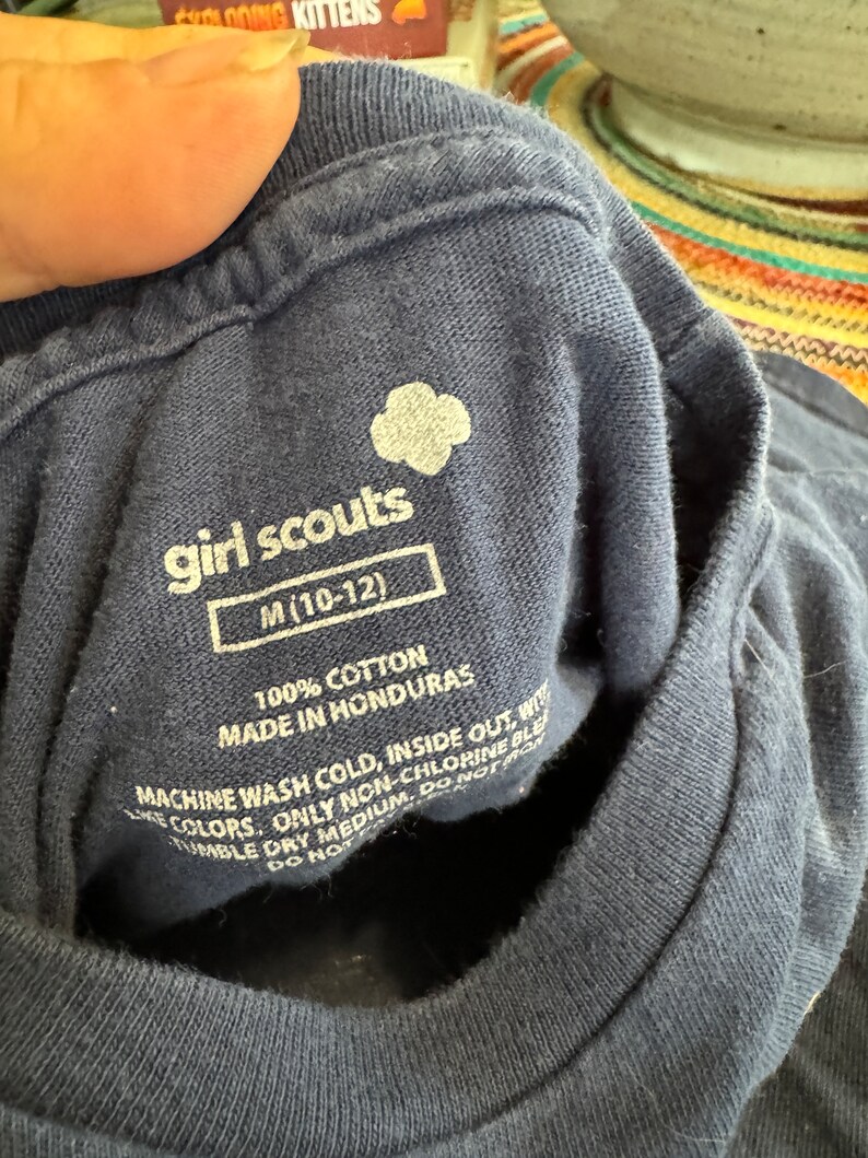 May include: A navy blue Girl Scouts t-shirt with the Girl Scouts logo and the text "girl scouts" and "M (10-12)". The tag also includes care instructions: "100% Cotton, Made in Honduras, Machine Wash Cold, Inside Out, With Like Colors, Only Non-Chlorine Bleach, Tumble Dry Medium, Do Not Iron, Do Not Dry Clean."