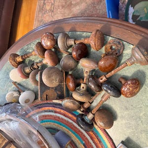May include: A collection of vintage door knobs in various shades of brown, with some having a ceramic finish. The knobs are arranged on a table with a green and brown surface.
