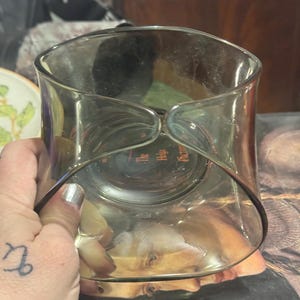 May include: A clear glass vase with a unique curved design. The vase has a wide, open top and a slightly narrower base. The sides curve inward, creating an hourglass-like shape. The vase is held by a hand with a tattoo.