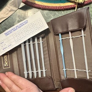 May include: A brown Sears crochet hook set with multiple gray crochet hooks and a blue crochet hook. The set includes a chart with crochet hook size equivalents. The case is open, revealing the hooks and a white chart.