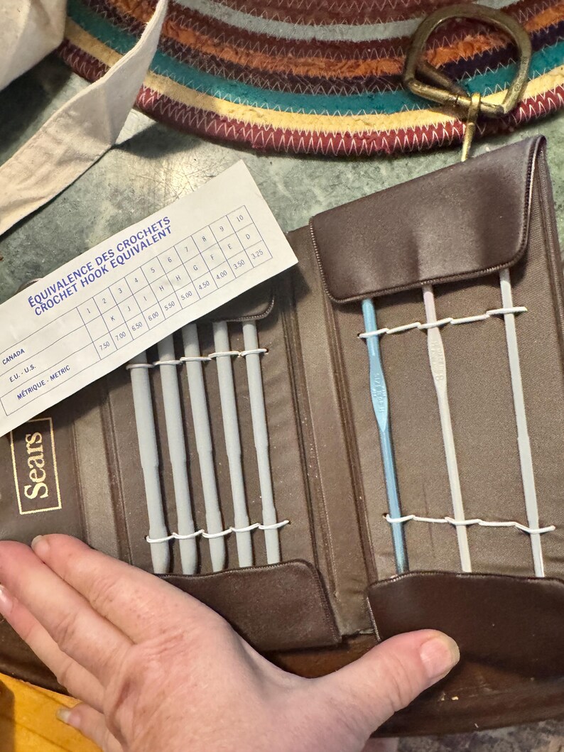 May include: A brown Sears crochet hook set with multiple gray crochet hooks and a blue crochet hook. The set includes a chart with crochet hook size equivalents. The case is open, revealing the hooks and a white chart.
