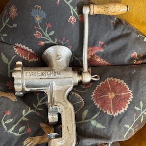 May include: A vintage, silver-colored meat grinder with a wooden handle. The grinder is marked with the number "5" and the text "MADE IN CHINA". It has a clamp for securing it to a surface.