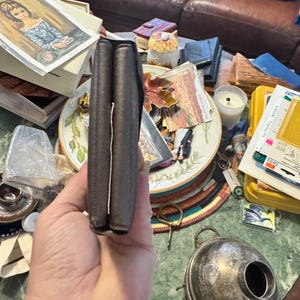 May include: A dark brown leather wallet, folded closed, held in a hand. The wallet appears to be made of a textured leather material. The edges are neatly stitched. The wallet is the primary focus, with a cluttered background.