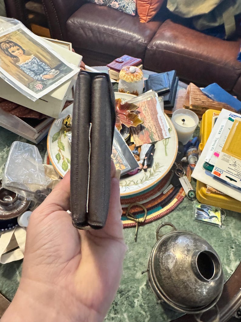 May include: A dark brown leather wallet, folded closed, held in a hand. The wallet appears to be made of a textured leather material. The edges are neatly stitched. The wallet is the primary focus, with a cluttered background.