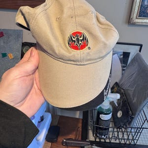 May include: Tan baseball cap with a black brim. The cap features a red, black, and white logo on the front. The brim has the word "Bacardi" printed on it. The cap is made of a soft material.