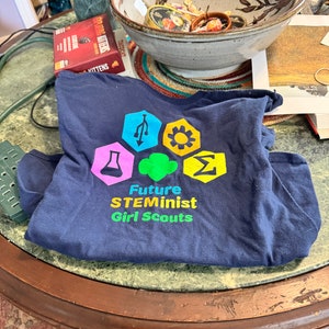 May include: A navy blue t-shirt with a colorful logo that says "Future STEMinist Girl Scouts". The logo features a variety of symbols, including a beaker, a gear, a lightbulb, and a computer chip.