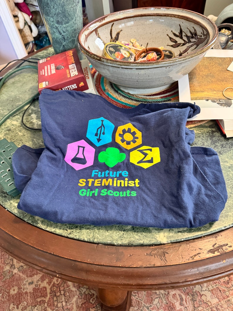 May include: A navy blue t-shirt with a colorful logo that says "Future STEMinist Girl Scouts". The logo features a variety of symbols, including a beaker, a gear, a lightbulb, and a computer chip.