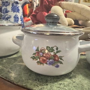 May include: White enamel cooking pot with a glass lid and side handles. The pot features a fruit basket design with red, purple, and green accents. A matching set of pots are visible on a green marble surface.
