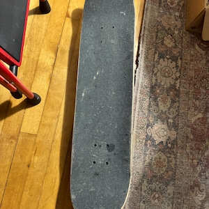 May include: A well-used skateboard with a black grip tape surface. The deck shows signs of wear and tear, with visible scuffs and marks. The skateboard is positioned on a wooden floor, with a portion of a red step stool visible in the background.