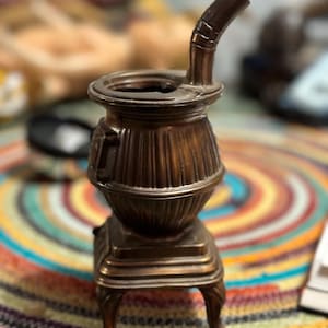 May include: A small, antique-looking metal stove with a decorative spout. The stove is brown and has a detailed design. It is sitting on a colorful rug.