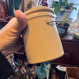 May include: A tan-colored, cylindrical stoneware crock with a matching lid and a metal handle. The crock has a slightly textured surface and a simple, classic design. It is a vintage kitchenware item.