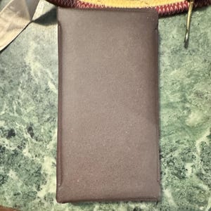 May include: A rectangular, brown case with a textured surface. The case is likely made of a soft material and has a darker brown trim. The case is sitting on a green and white marble-patterned surface.