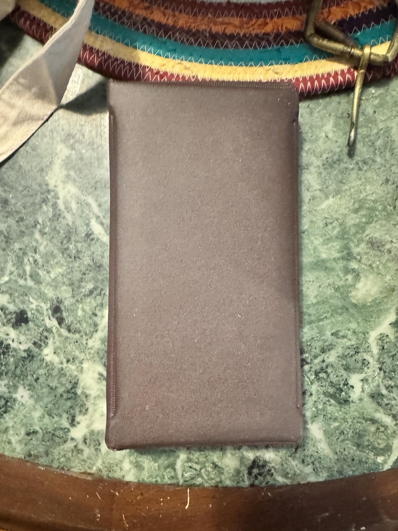 May include: A rectangular, brown case with a textured surface. The case is likely made of a soft material and has a darker brown trim. The case is sitting on a green and white marble-patterned surface.