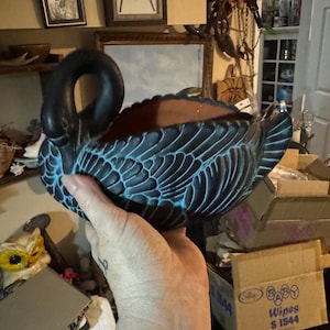 May include: A swan-shaped ceramic planter featuring a matte black finish with teal blue highlights accentuating the molded feather details. The interior of the vessel is a smooth, unglazed terracotta brown color, and the swan has a curved neck.