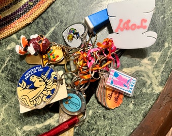 May include: A collection of keychains with various designs, including a white ceramic cup with the word "Kool" in pink lettering, a colorful unicorn keychain with the words "Niagara Falls, NY", a red and blue keychain with a cartoon character, and a keychain with a butterfly design. The keychains are attached to a silver key ring and are on a green marble surface.