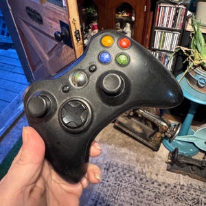 May include: A black wireless video game controller featuring a central circular guide button, two analog sticks, a directional pad, and four colored action buttons in yellow, red, blue, and green. The controller includes small start and back buttons.