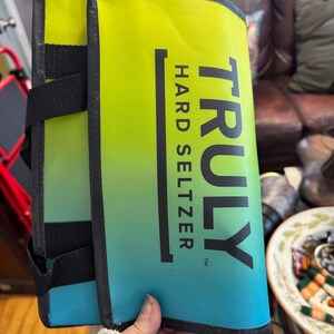 May include: A folded, insulated beverage carrier with a gradient design of yellow, green, and blue. The carrier features the text "TRULY HARD SELTZER" in black. Black straps and trim are visible.