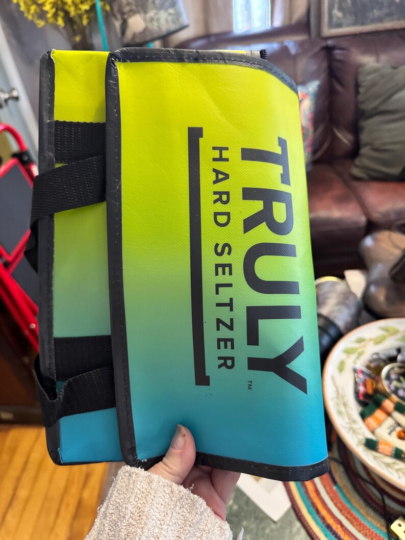 May include: A folded, insulated beverage carrier with a gradient design of yellow, green, and blue. The carrier features the text "TRULY HARD SELTZER" in black. Black straps and trim are visible.