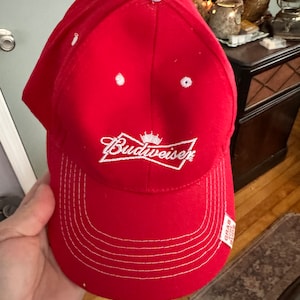 May include: Red baseball cap with a white Budweiser logo and white stitching. The cap has a white tag that says "Grab Some Buds".