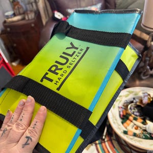 May include: A rectangular, insulated cooler bag with the text "TRULY HARD SELTZER" in black. The bag features a gradient of yellow and green, with a blue top and black straps. The bag is designed to keep beverages cold.