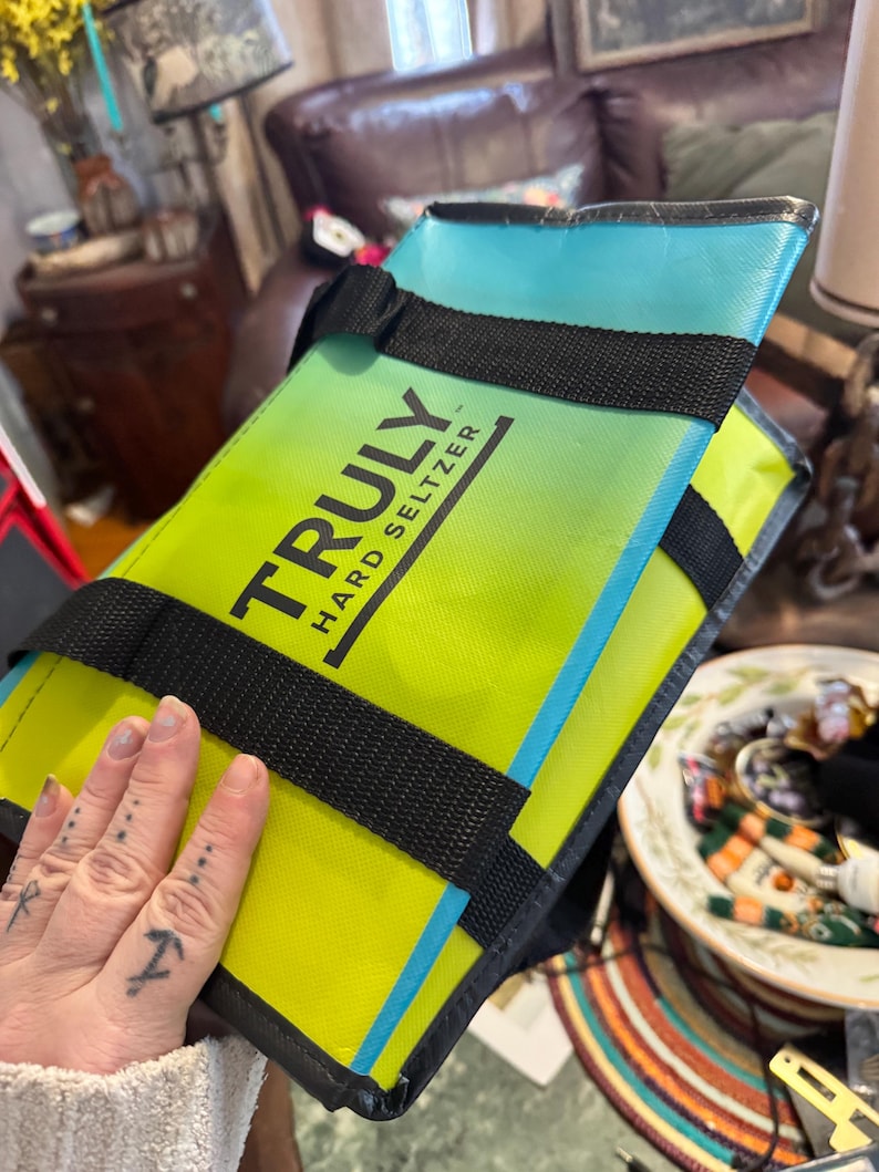 May include: A rectangular, insulated cooler bag with the text "TRULY HARD SELTZER" in black. The bag features a gradient of yellow and green, with a blue top and black straps. The bag is designed to keep beverages cold.