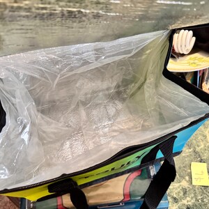 May include: Open insulated tote bag with a black zipper and handles. The bag has a blue, green, and yellow exterior with a silver interior lining. The bag is open, revealing the interior lining.
