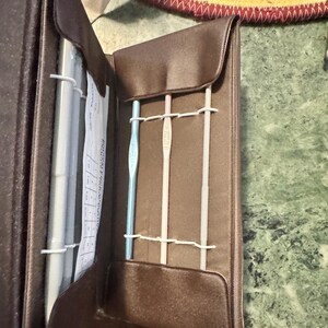 May include: A brown, open crochet hook case with three crochet hooks secured by white elastic bands. The case is made of a smooth, leather-like material. A paper with text is visible inside the case.
