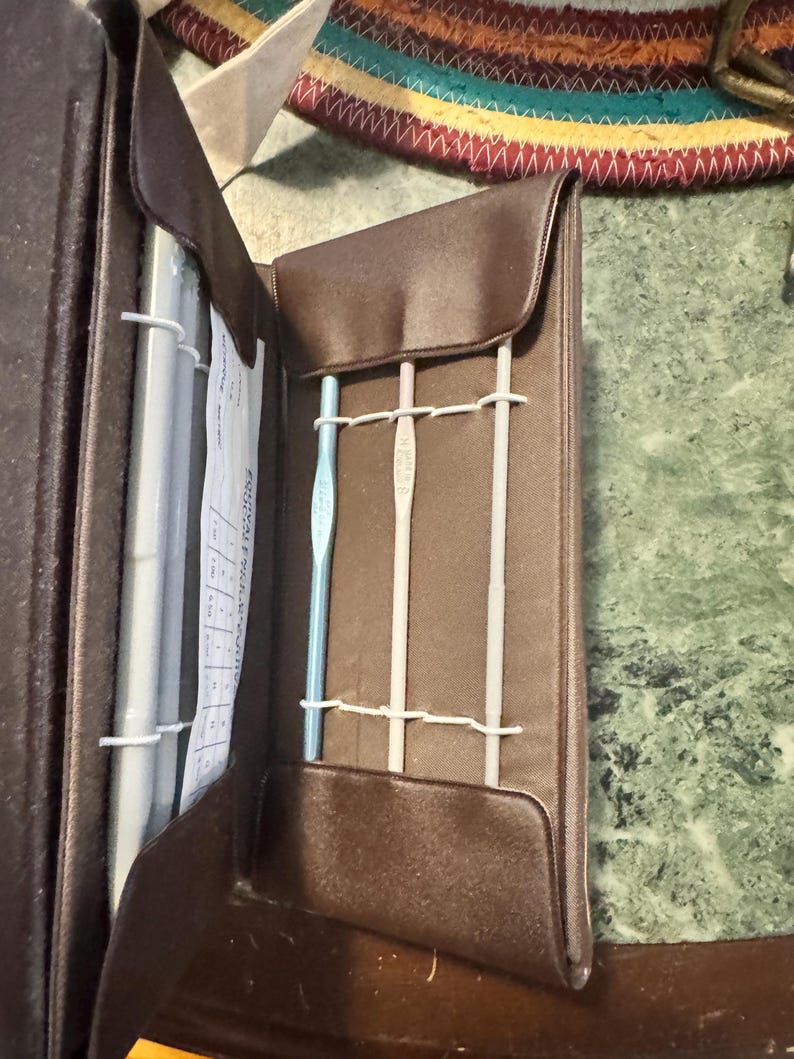May include: A brown, open crochet hook case with three crochet hooks secured by white elastic bands. The case is made of a smooth, leather-like material. A paper with text is visible inside the case.