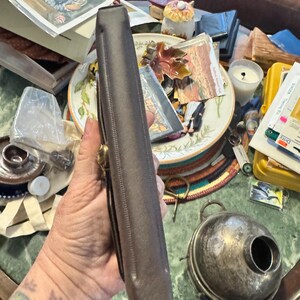 May include: A brown leather case with a gold-colored clasp. The case is rectangular and appears to be closed. The image also shows a silver-colored container with a round body and a handle.