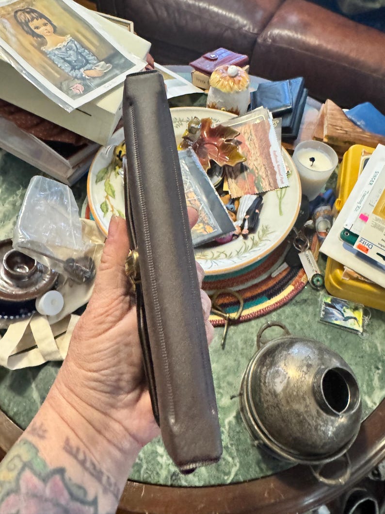 May include: A brown leather case with a gold-colored clasp. The case is rectangular and appears to be closed. The image also shows a silver-colored container with a round body and a handle.