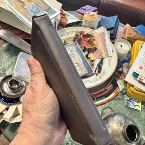 May include: A dark brown, rectangular leather case with a stitched seam. The case is held at an angle, revealing its shape and texture. The background includes various items on a table, suggesting a vintage or antique theme.