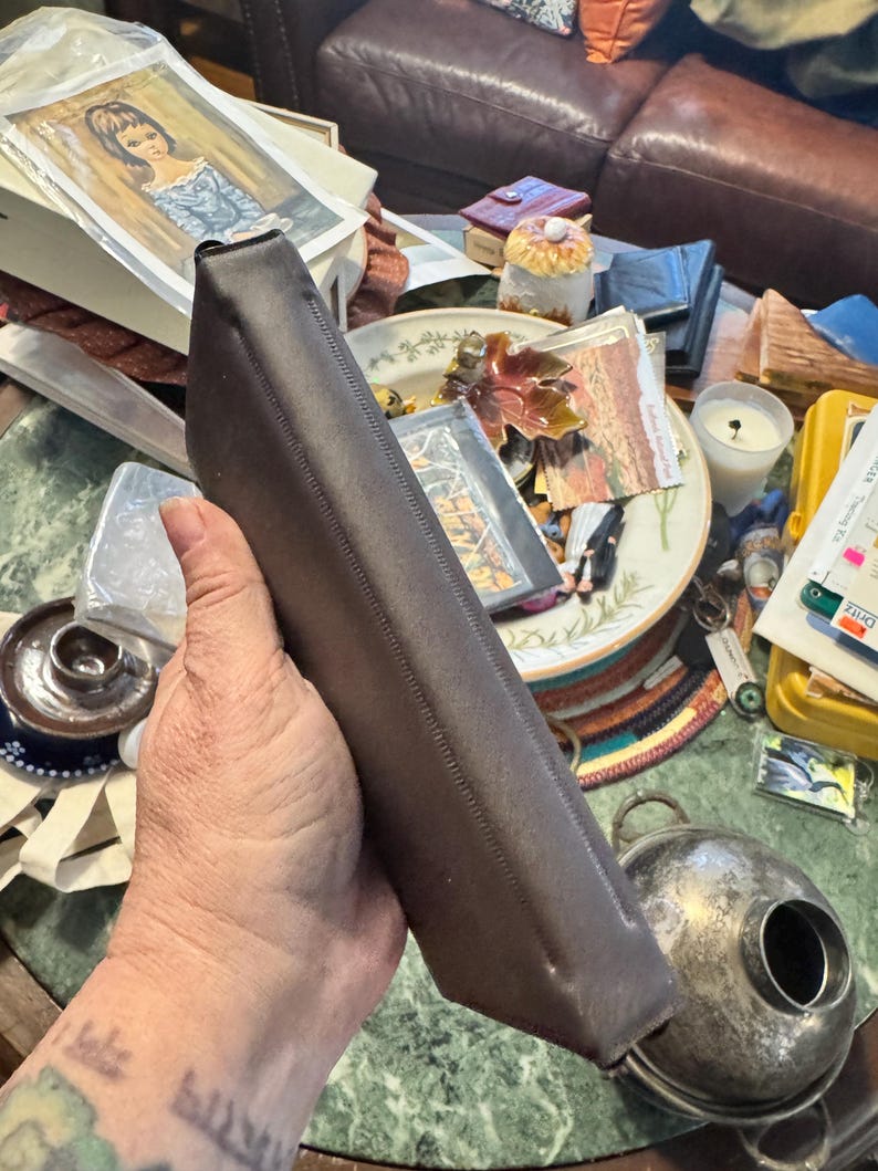 May include: A dark brown, rectangular leather case with a stitched seam. The case is held at an angle, revealing its shape and texture. The background includes various items on a table, suggesting a vintage or antique theme.