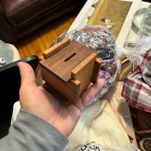 May include: A small wooden box with a slot on the top. The box is made of dark wood with light wood trim. The box is being held in a person's hand.
