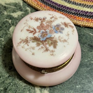 May include: A round, pink trinket box with a floral design on the lid. The lid features a delicate illustration of blue and brown flowers and foliage. The box has a gold-colored metal band around the middle and sits on a green marble surface.