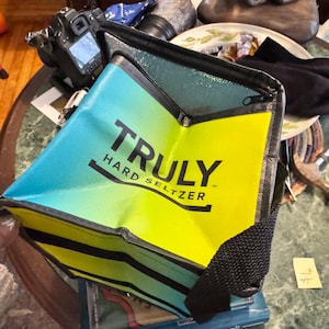May include: A blue, yellow, and black insulated cooler bag with the text "TRULY HARD SELTZER" printed on the front. The bag has a black strap and is partially open, revealing the interior.