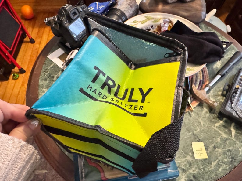 May include: A blue, yellow, and black insulated cooler bag with the text "TRULY HARD SELTZER" printed on the front. The bag has a black strap and is partially open, revealing the interior.