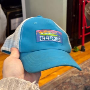 May include: A blue baseball cap with a white mesh back. The cap features the LLBean logo on the front, with a colorful mountain and sunset design. The brim is curved and the cap is made of a durable material.