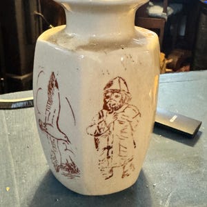 May include: Cream-colored ceramic vase with a flared rim and a hexagonal body. The vase features brown illustrations of a bird in flight and a person in a hat and coat. The vase has a rustic, vintage aesthetic.