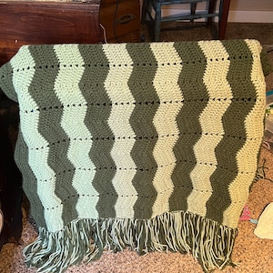May include: A crocheted blanket with a wavy pattern in shades of green. The blanket has a fringe border.